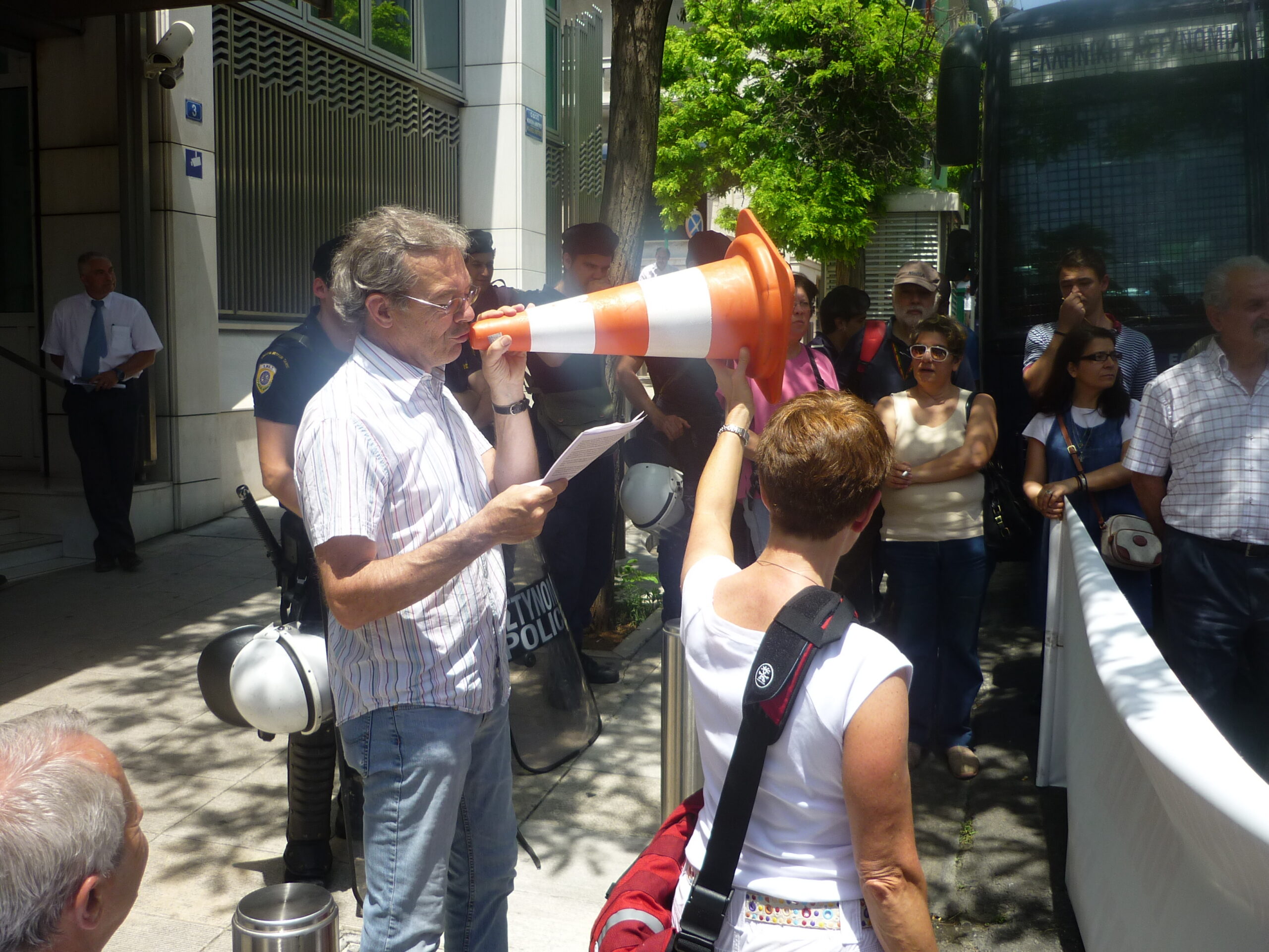 Achim Rollhäuser spricht durch ein Baustellenhütchen zu der Kundgebung vor der deutschen Botschaft in Athen